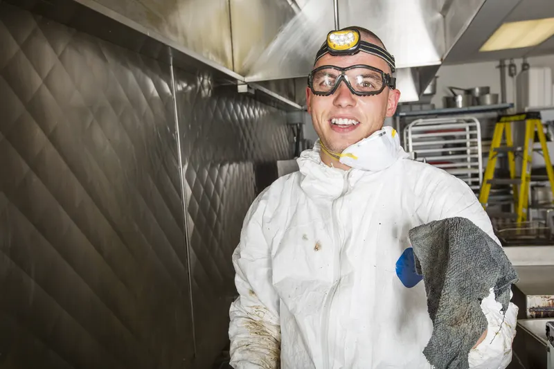 Hood cleaning technician in white coveralls and a headlamp holding a cleaning cloth beside a stainless hood