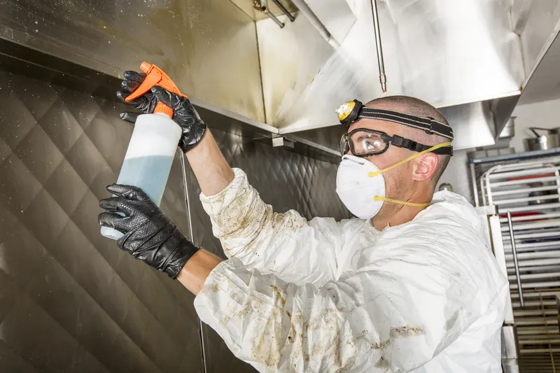 Technician in gloves and a respirator spraying cleaner up under a stainless restaurant hood